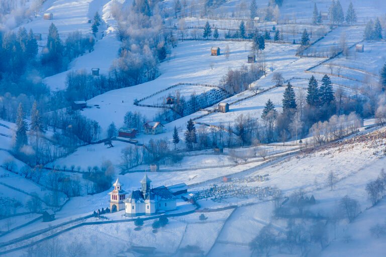 Peisaj de iarnă cu biserică și dealuri înzăpezite.