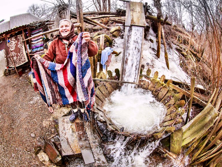 Femeie ținând covoare colorate lângă apă curgătoare.