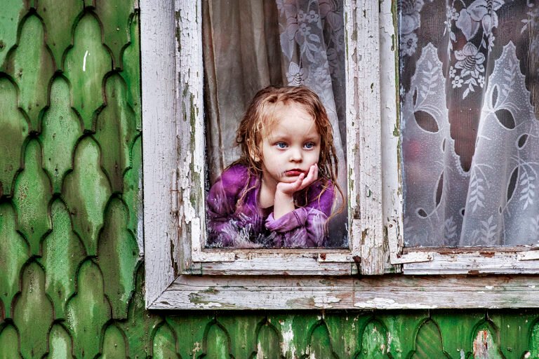 Fetiță privind gânditoare pe fereastra unei case verzi.