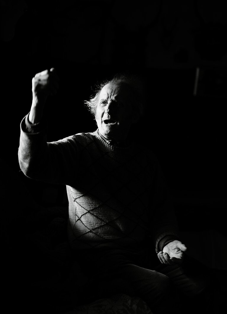 Bătrân gesticulând dramatic în lumină slabă.