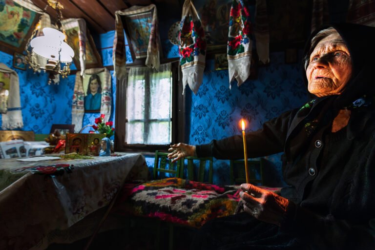 Bătrână cu lumânare într-o cameră tradițională românească.