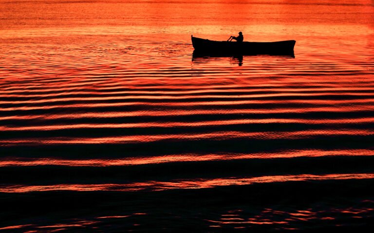 Canoeist la apus pe apă cu reflexii roșii.